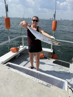 Person on a boat holding a large fish with water and city skyline in the background on a sunny day.