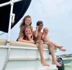Four children sit on the edge of a boat, smiling at the camera with water and distant buildings in the background.