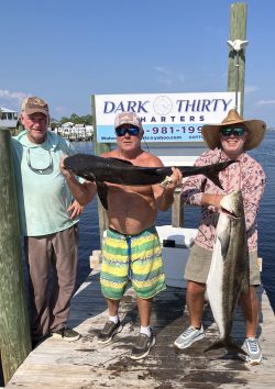 Screenshot Three men on a dock hold large fish in front of a Dark Thirty Charters sign on a sunny day.