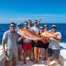 A group of people on a boat hold up two large fish, smiling, with the ocean and blue sky in the background.