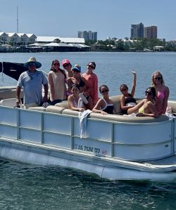 Screenshot A group of people pose on a pontoon boat in the water with buildings and blue sky visible in the background.