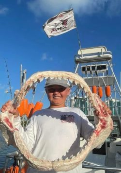 Smiling child holding a large shark jaw on a boat, with fishing gear and a flag visible in the background.
