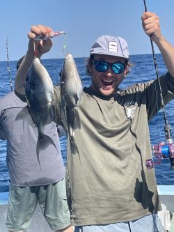 Man on a boat holding up two fish with one hand, smiling, with another person fishing in the background.